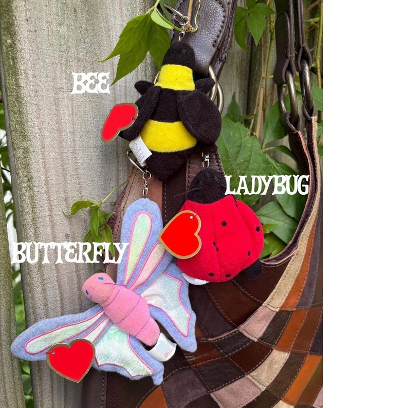 K&ouml;nnte beinhalten: Drei Pl&uuml;sch-Schl&uuml;sselanh&auml;nger in Form von Insekten und Tieren an einer braunen Ledertasche. Die Artikel umfassen eine gelb-schwarze Biene, einen roten Marienk&auml;fer mit schwarzen Punkten und einen pastellfarbenen rosa-blauen Schmetterling mit Herz.
