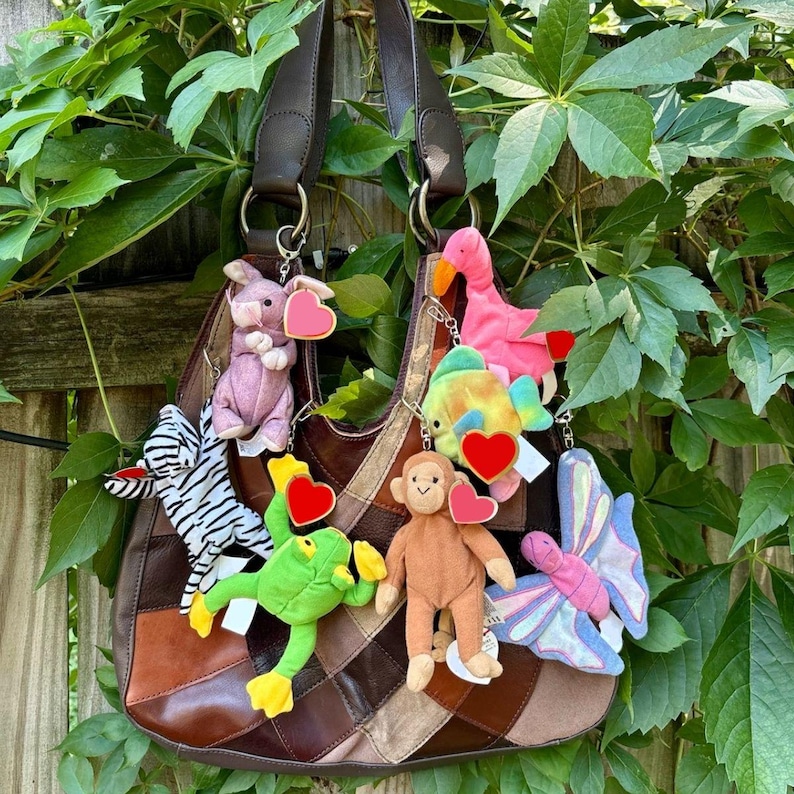 K&ouml;nnte beinhalten: Eine braune Patchwork-Ledertasche, die mit mehreren Pl&uuml;schtier-Schl&uuml;sselanh&auml;ngern verziert ist, darunter ein lila Schwein, ein Zebra, ein gr&uuml;ner Frosch, ein rosa Flamingo, ein bunter Fisch, ein brauner Affe und ein Schmetterling mit Herzanh&auml;ngern.