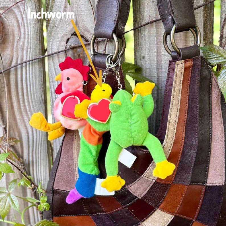K&ouml;nnte beinhalten: Eine braune Leder-Patchwork-Handtasche mit drei Pl&uuml;sch-Schl&uuml;sselanh&auml;ngern an den Metallringen. Die Anh&auml;nger zeigen ein rosa Huhn, einen gelben und regenbogenfarbenen Wurm mit dem Text Inchworm sowie einen gr&uuml;nen Frosch, jeweils mit einem roten Herzanh&auml;nger.