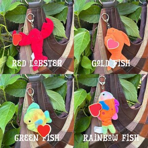 K&ouml;nnte beinhalten: Eine Sammlung von vier Pl&uuml;sch-Schl&uuml;sselanh&auml;ngern, bestehend aus einem roten Hummer, einem orangefarbenen Goldfisch, einem gr&uuml;nen Fisch und einem bunten Regenbogenfisch. Jedes Pl&uuml;schtier hat einen silbernen Metallring und ein rotes herzf&ouml;rmiges Etikett.