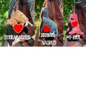 K&ouml;nnte beinhalten: Drei Dinosaurier-Pl&uuml;sch-Schl&uuml;sselanh&auml;nger an einer braunen Ledertasche. Der beige Stegosaurus, der blaue Brontasaurus und der rosa T-Rex haben jeweils ein rotes herzf&ouml;rmiges Etikett, wobei der Name des Dinosauriers in Wei&szlig; darunter steht.