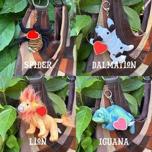 K&ouml;nnte beinhalten: Eine Sammlung von vier Pl&uuml;schtier-Schl&uuml;sselanh&auml;ngern an einer braunen Patchwork-Ledertasche. Das Set enth&auml;lt eine schwarze und orange Spinne, einen wei&szlig;-schwarzen Dalmatiner, einen gelb-orangen L&ouml;wen und einen blau-gr&uuml;nen Leguan, jeweils mit einem Herzanh&auml;nger.