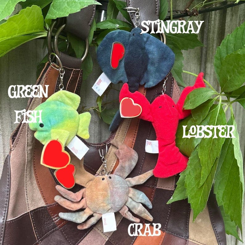 K&ouml;nnte beinhalten: Eine Sammlung von vier Pl&uuml;sch-Tier-Schl&uuml;sselanh&auml;ngern, bestehend aus einem gr&uuml;nen Fisch, einem dunkelblauen Stachelrochen, einem leuchtend roten Hummer und einer beigen Krabbe. Jedes Pl&uuml;schtier hat ein rotes herzf&ouml;rmiges Etikett und einen Metallring.