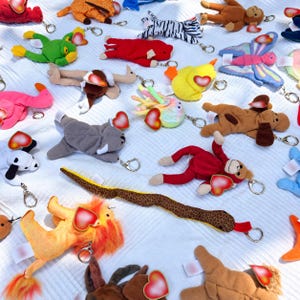 K&ouml;nnte beinhalten: Eine Sammlung verschiedener Pl&uuml;schtier-Schl&uuml;sselanh&auml;nger mit kleinen Stofffiguren wie einem L&ouml;wen, einer Schlange, einem Affen, einem Frosch und einem Schmetterling. Jedes Stofftier ist an einem Metallring befestigt und hat ein rotes Herz-Etikett.