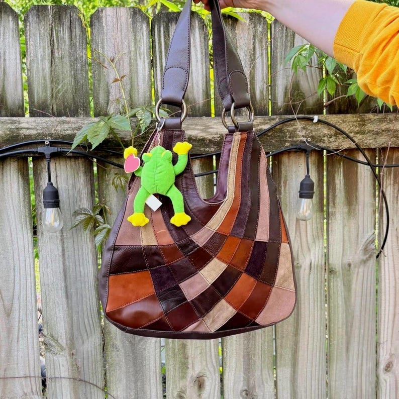 K&ouml;nnte beinhalten: Eine Hobo-Schultertasche aus mehrfarbigen Lederflicken in Braun-, Beige- und Cremet&ouml;nen, die in einem f&auml;cherf&ouml;rmigen Muster angeordnet sind. Die Tasche verf&uuml;gt &uuml;ber einen einzelnen Schulterriemen und einen hellgr&uuml;nen Pl&uuml;schfrosch-Anh&auml;nger mit einem rosa Herz.