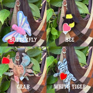 K&ouml;nnte beinhalten: Eine Sammlung von vier Pl&uuml;schtier-Schl&uuml;sselanh&auml;ngern, bestehend aus einem rosa-blauen Schmetterling, einer schwarz-gelben Biene, einer beigen Krabbe und einem wei&szlig;-schwarz gestreiften Tiger. Jedes Pl&uuml;schtier hat ein herzf&ouml;rmiges Etikett und einen Metallring.