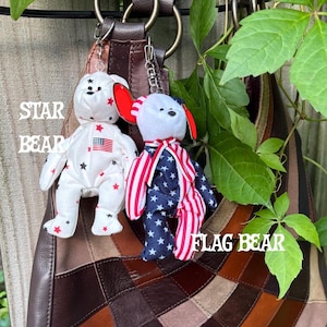 K&ouml;nnte beinhalten: Zwei Pl&uuml;schb&auml;ren-Schl&uuml;sselanh&auml;nger an einer braunen Ledertasche. Der linke B&auml;r ist wei&szlig; mit roten und schwarzen Sternen und einem kleinen amerikanischen Flaggenaufn&auml;her. Der rechte B&auml;r hat einen wei&szlig;en Kopf, ein rot-wei&szlig; gestreiftes Halsband und einen K&ouml;rper mit blau-wei&szlig;en Sternen sowie rot-wei&szlig;en Streifen.