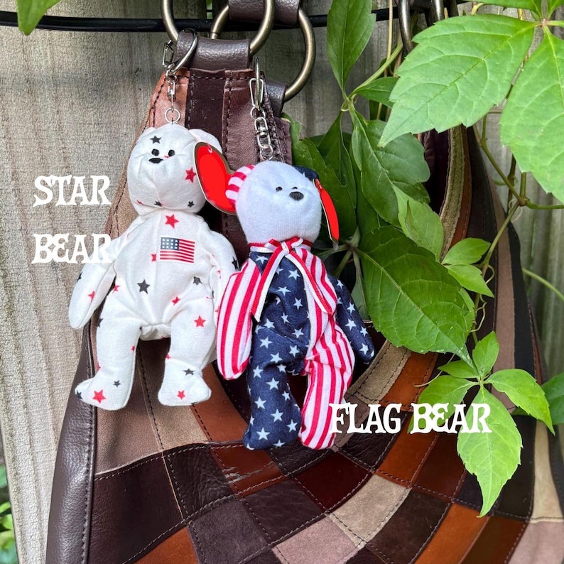 K&ouml;nnte beinhalten: Zwei Pl&uuml;schb&auml;ren-Schl&uuml;sselanh&auml;nger an einer braunen Ledertasche. Der linke B&auml;r ist wei&szlig; mit roten und schwarzen Sternen und einem kleinen amerikanischen Flaggenaufn&auml;her. Der rechte B&auml;r hat einen wei&szlig;en Kopf, ein rot-wei&szlig; gestreiftes Halsband und einen K&ouml;rper mit blau-wei&szlig;en Sternen sowie rot-wei&szlig;en Streifen.