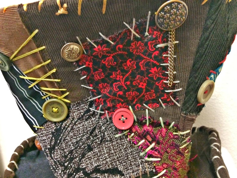 May include: A patchwork textile composed of diverse fabrics like corduroy, patterned red and black fabric, and tweed. It features visible stitching, buttons, and a decorative chain. The color scheme includes brown, red, black, and beige.