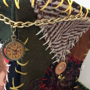 May include: A patchwork fabric item with a gold chain and a clock charm. The item features various fabrics in green, brown, and patterned textiles. A gold hook is attached to the side. Decorative buttons and stitching are visible.