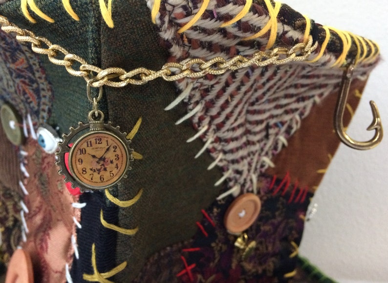 May include: A patchwork fabric item with a gold chain and a clock charm. The item features various fabrics in green, brown, and patterned textiles. A gold hook is attached to the side. Decorative buttons and stitching are visible.