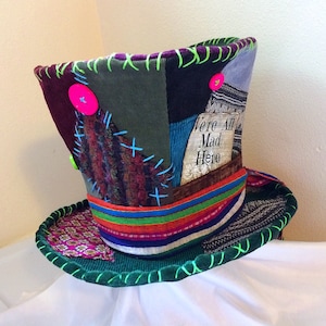 May include: A patchwork top hat with a wide brim, featuring various fabrics in shades of green, purple, and blue. The hat is adorned with neon green stitching, a pink button, and the text "We're All Mad Here". A whimsical accessory.