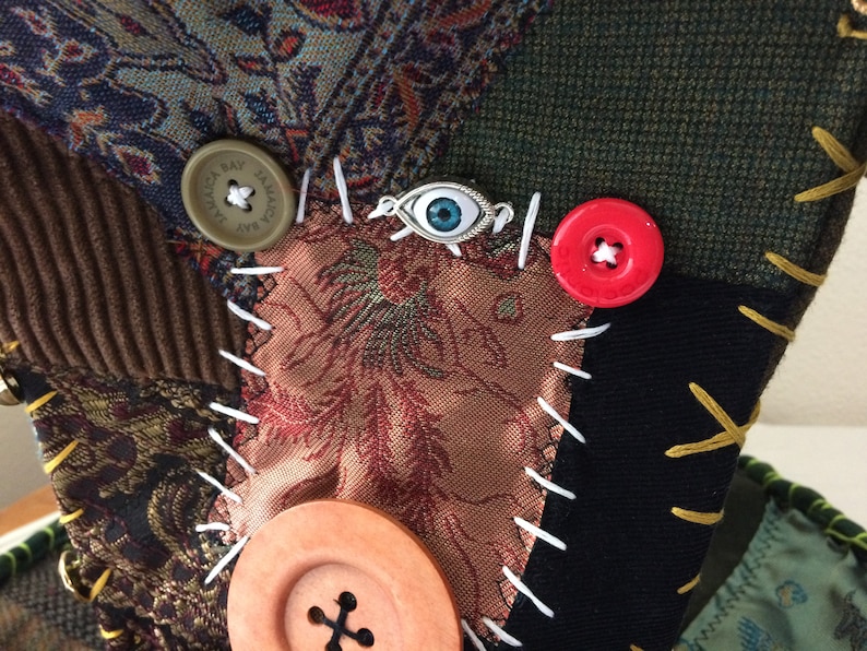 May include: Close-up of a patchwork textile with various fabric patterns and textures. Features a brown corduroy section, floral print, and a dark green section. Adorned with buttons in brown and red, and a silver eye charm. White and yellow stitching.