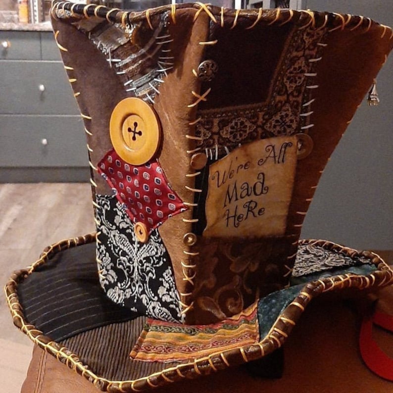 May include: A brown top hat in the style of the Mad Hatter, featuring a wide brim and patchwork design. The hat incorporates various fabrics, wooden buttons, and decorative stitching. The words "We're All Mad Here" are written on the side.
