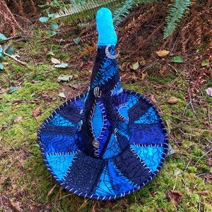 May include: A blue and black witch hat with a pointed crown and a crescent moon charm. The hat is made of patchwork fabric with a spiderweb design.