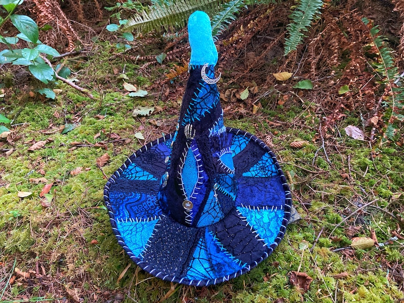 May include: A blue and black witch hat with a pointed crown and a crescent moon charm. The hat is made of patchwork fabric with a spiderweb design.