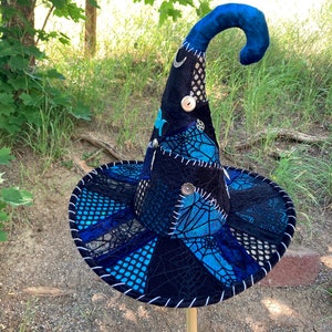 May include: A black and blue witch hat with a pointed crown and a curled brim. The hat is decorated with silver accents, including a crescent moon, a star, and a circle with a cross inside. The hat is made of a variety of fabrics, including velvet, lace, and mesh.