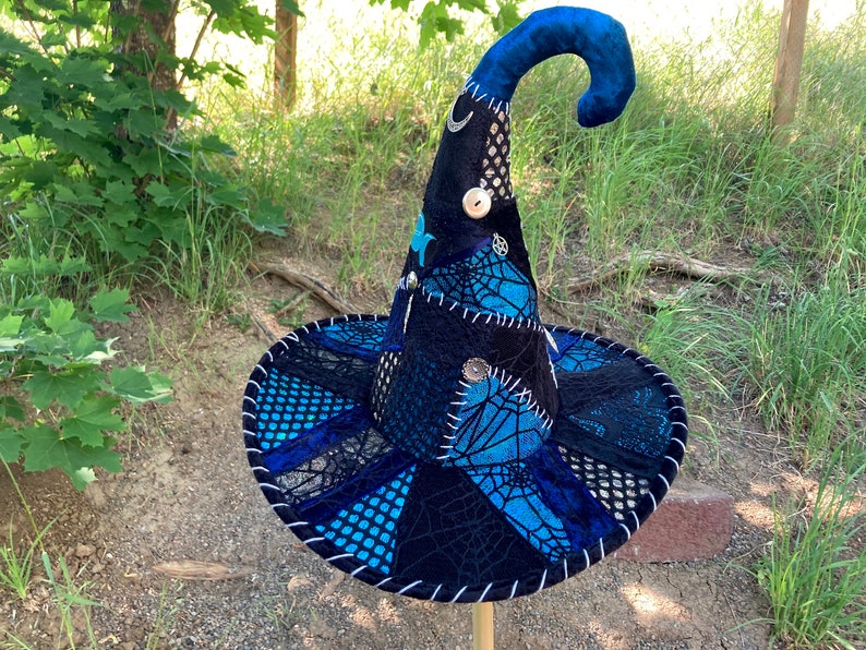 May include: A black and blue witch hat with a pointed crown and a curled brim. The hat is decorated with silver accents, including a crescent moon, a star, and a circle with a cross inside. The hat is made of a variety of fabrics, including velvet, lace, and mesh.