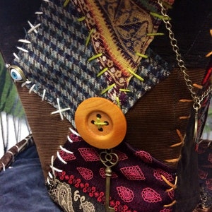 May include: A patchwork hat with a variety of fabrics, including tweed, paisley, and velvet. The hat features a large wooden button, a decorative key, and is embellished with contrasting stitching. The colour palette includes brown, blue, burgundy, and gold.