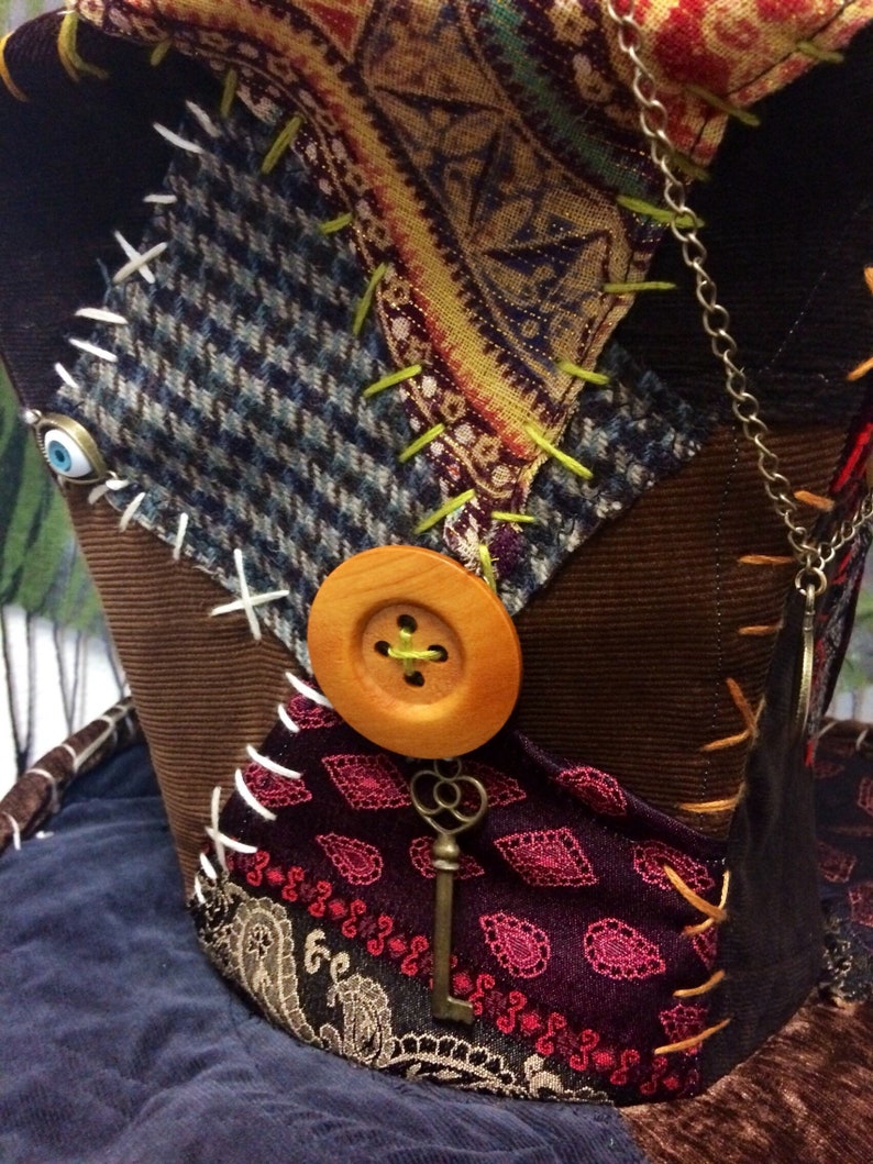 May include: A patchwork hat with a variety of fabrics, including tweed, paisley, and velvet. The hat features a large wooden button, a decorative key, and is embellished with contrasting stitching. The colour palette includes brown, blue, burgundy, and gold.