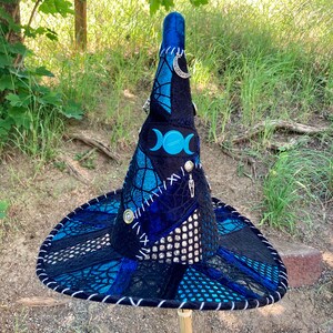 May include: A blue and black witch hat with a pointed crown and a wide brim. The hat is decorated with a triple moon symbol, a skeleton charm, and a crescent moon charm. The hat is made of a variety of fabrics, including velvet, lace, and mesh.