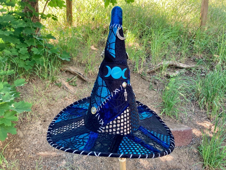 May include: A blue and black witch hat with a pointed crown and a wide brim. The hat is decorated with a triple moon symbol, a skeleton charm, and a crescent moon charm. The hat is made of a variety of fabrics, including velvet, lace, and mesh.