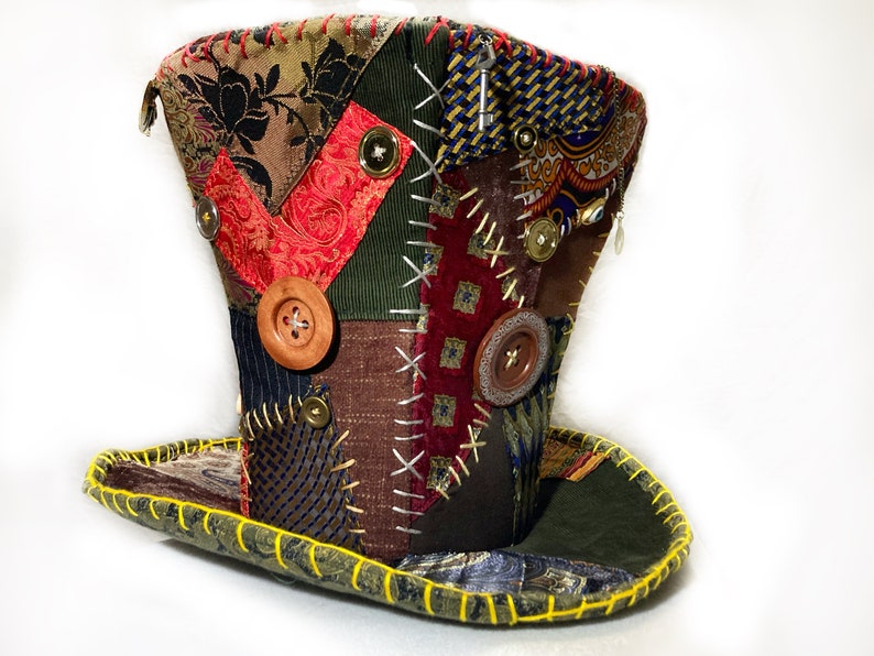 May include: A patchwork top hat with a wide brim, showcasing diverse fabric patterns and colours, such as floral, paisley, and striped designs. The hat is embellished with wooden buttons and decorative stitching, with a yellow stitched brim.
