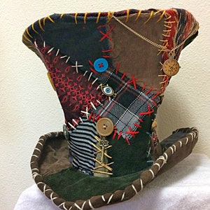 May include: A patchwork top hat featuring a mix of fabrics like plaid, stripes, and corduroy. The hat is decorated with buttons, charms, and red, yellow, and white stitching. A gold chain with a small clock pendant hangs from the side.