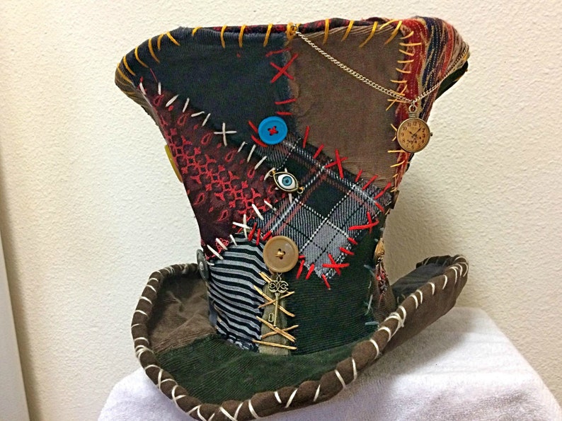 May include: A patchwork top hat featuring a mix of fabrics like plaid, stripes, and corduroy. The hat is decorated with buttons, charms, and red, yellow, and white stitching. A gold chain with a small clock pendant hangs from the side.
