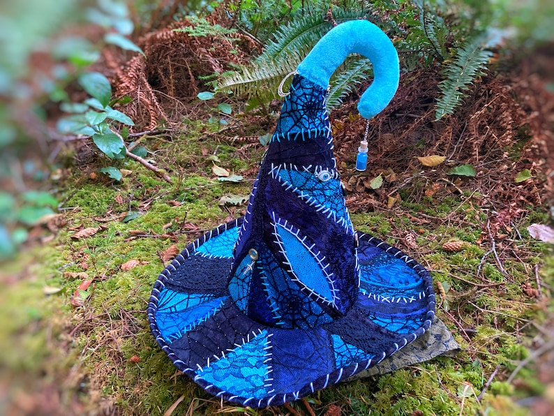 May include: A blue and black witch hat with a pointed crown and a curled brim. The hat is decorated with a spiderweb design and a small blue bottle charm.