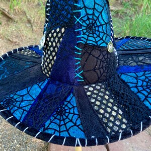 May include: A wide-brimmed hat with a patchwork design featuring black, blue, and silver fabric. The hat is decorated with a spiderweb pattern and has a white trim around the edge.