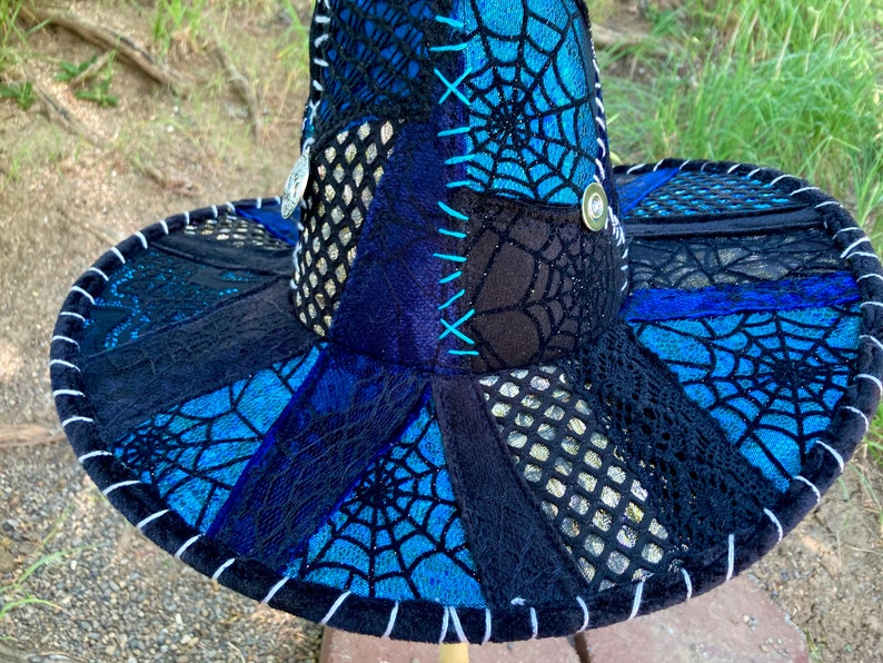 May include: A wide-brimmed hat with a patchwork design featuring black, blue, and silver fabric. The hat is decorated with a spiderweb pattern and has a white trim around the edge.