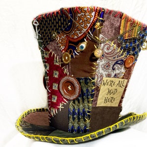 Pode incluir: Um chapéu de copa de patchwork com aba larga, apresentando vários padrões e texturas de tecido. Adornado com botões, amuletos e o texto "WE'RE ALL MAD HERE". A aba do chapéu é delineada com costura amarela, criando um acessório caprichoso.