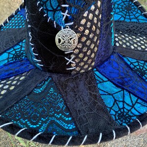 May include: A black wide-brimmed hat with a blue and silver patterned design. The hat is made of different fabrics and has a white stitching detail around the brim. A silver charm with a tree of life design is attached to the hat.