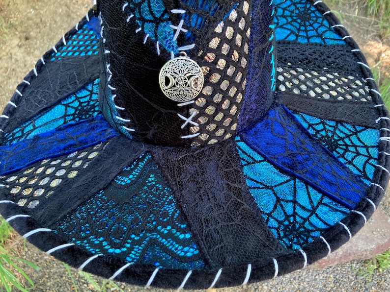 May include: A black wide-brimmed hat with a blue and silver patterned design. The hat is made of different fabrics and has a white stitching detail around the brim. A silver charm with a tree of life design is attached to the hat.
