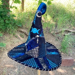 May include: A blue and black witch hat with a pointed crown, a curled brim, and a variety of embellishments, including a triple moon pendant, a crescent moon charm, and a silver skeleton charm.