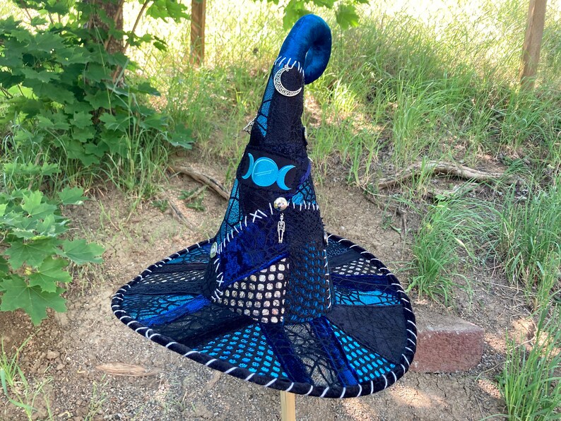 May include: A blue and black witch hat with a pointed crown, a curled brim, and a variety of embellishments, including a triple moon pendant, a crescent moon charm, and a silver skeleton charm.