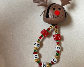May include: A brown reindeer bell ornament with a red nose and a white beaded chain with red dice and white letter beads spelling "UNCOCO".