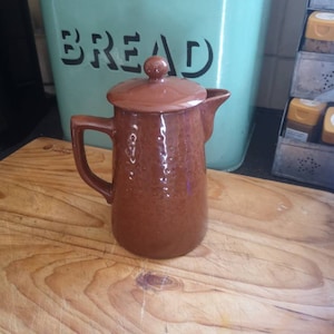 May include: A brown ceramic coffee pot with a lid and a handle. The pot is sitting on a wooden surface.