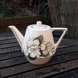May include: White ceramic teapot with a floral design featuring white flowers and green leaves. The teapot has a lid and a spout.