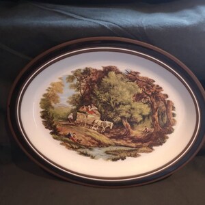 以下が含まれることがあります： 茶色の縁取りのある白い楕円形のセラミック製サービングプレート。プレートには、森の中を走る馬車のヴィンテージスタイルのイラストが描かれています。