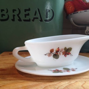 Puede incluir: Salsera de cerámica blanca con un diseño floral con bayas rojas y hojas verdes. La salsera está sobre un platillo blanco a juego.