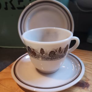 May include: A white ceramic teacup and saucer with a brown line design and a brown tree pattern. The cup has a handle and the saucer has a brown line around the edge.