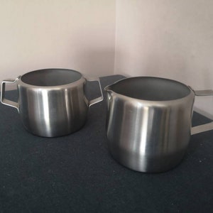 May include: Two stainless steel creamer pitchers, one with a handle on the right side and the other with handles on both sides.