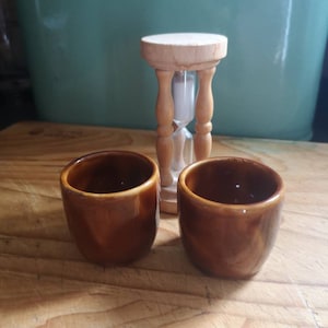 Puede incluir: Dos hueveras de cerámica marrón sobre una superficie de madera. Un reloj de arena está en el fondo.