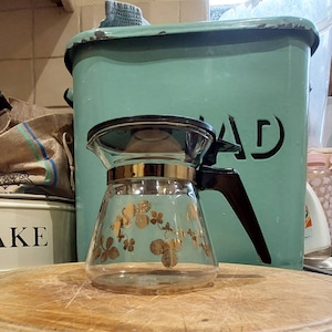 May include: A clear glass coffee pot with a black handle and gold clover leaf design. The pot is sitting on a wooden chopping board.