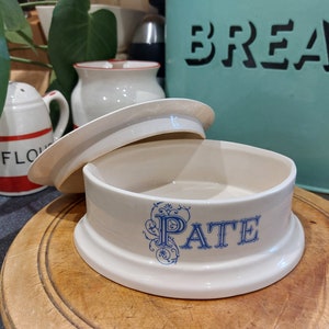 Könnte beinhalten: Weiße Keramik-Pate-Schale mit blau-weißem Blumendesign und dem Wort "PATE" in blauer Schrift. Die Schale hat einen Deckel und steht auf einem Holzbrett.