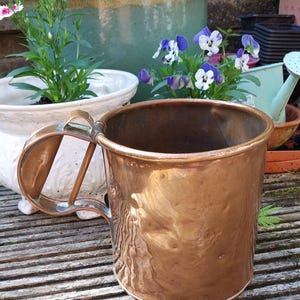 May include: A large, antique copper mug with a handle. The mug has a hammered texture and a shiny, aged finish. The mug is sitting on a wooden surface, with potted plants and flowers in the background.