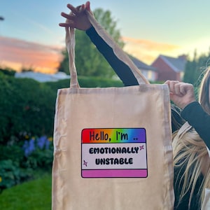 May include: A beige tote bag featuring the text "Hello, I'm... EMOTIONALLY UNSTABLE" in a colourful graphic. The bag is held up against a backdrop of green foliage and a blue sky.