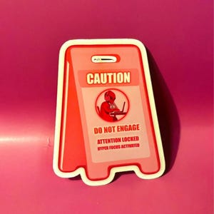 May include: A die-cut sticker shaped like a pink caution sign. The text reads CAUTION, DO NOT ENGAGE, ATTENTION LOCKED, HYPER FOCUS ACTIVATED. A circular graphic shows a silhouette of a person wearing headphones while working on a laptop computer.